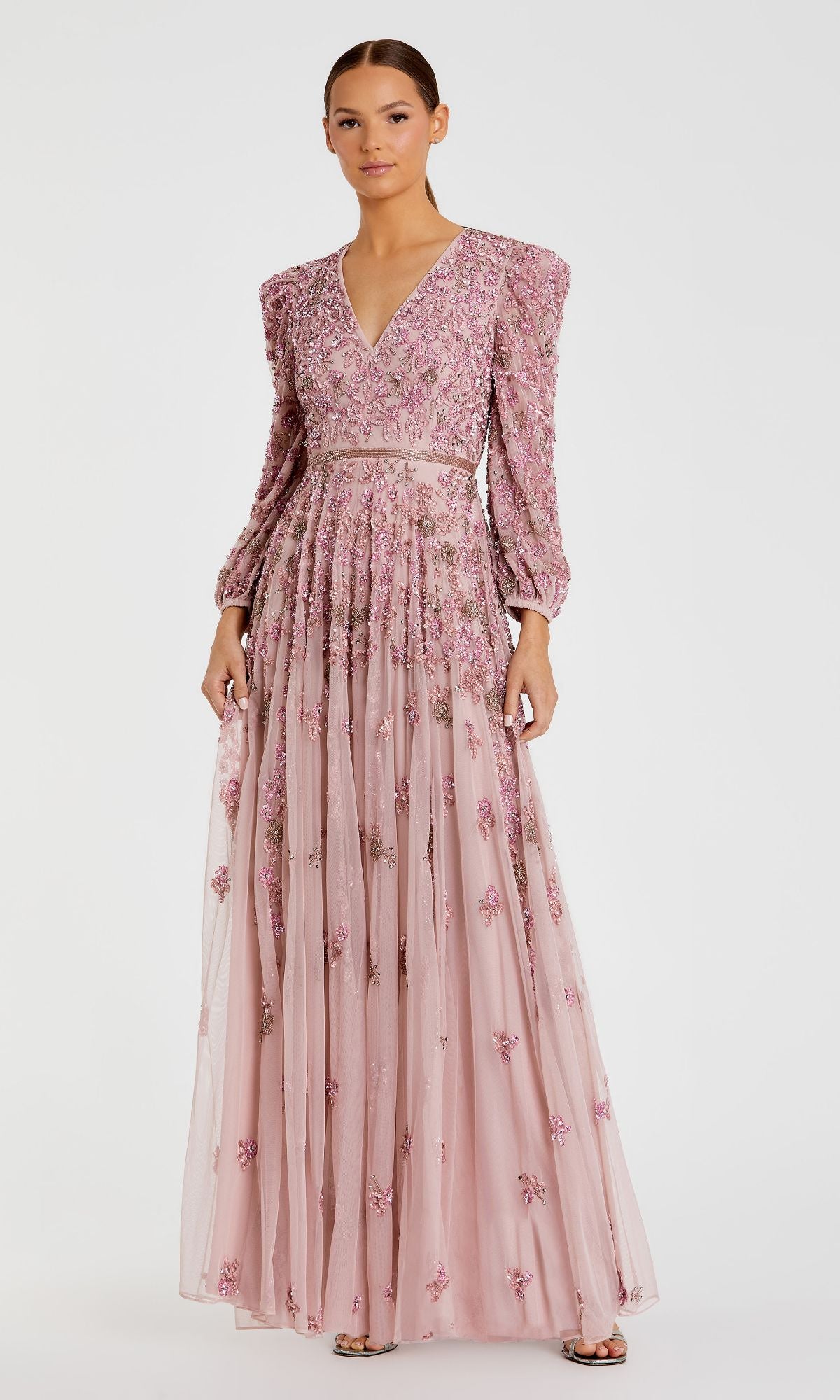 A person stands against a plain white background wearing the Mac Duggal 5497 Long Sleeve Beaded Formal Dress—a modest pink gown featuring floral embroidery, long sleeves, a V-neckline, and a fitted waist.